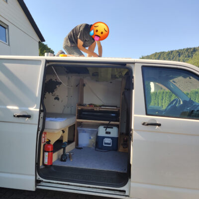 Ausbau: Auf dem Bild ist ein VW Transporter zu sehen. Eine Person kniet auf dem Dach und sägt ein Loch hinein.